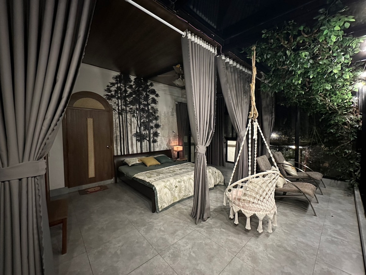 A spacious sleeping area features a large bed with decorative bedding, complemented by a wall adorned with a nature-inspired mural. A cozy hanging chair is positioned beside the bed, while elegant curtains frame the space and enhance the connection to the greenery visible outside.