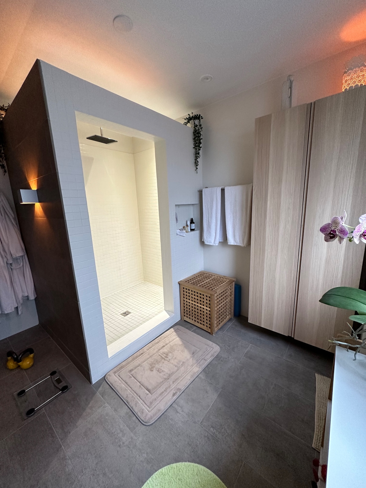 A modern shower area features a spacious walk-in design with white tiles and a rain showerhead. Adjacent are two towels hung neatly, a bamboo storage basket, and a small mat on the floor. Soft lighting creates a calming atmosphere.