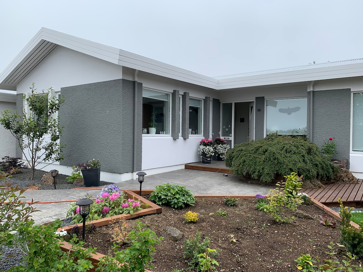 A welcoming exterior of the property is presented, showcasing a modern design with gray and white accents. A landscaped garden features colorful flower beds and a manicured shrub, while outdoor lighting fixtures are positioned along the path.