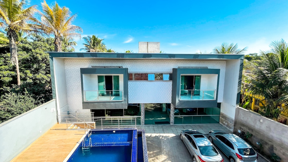 The modern, two-story building features a smooth, white exterior complemented by large glass balconies. A swimming pool is situated adjacent to the structure, with a patio area offering space for outdoor activities. Lush palm trees surround the property, enhancing the tropical ambiance.