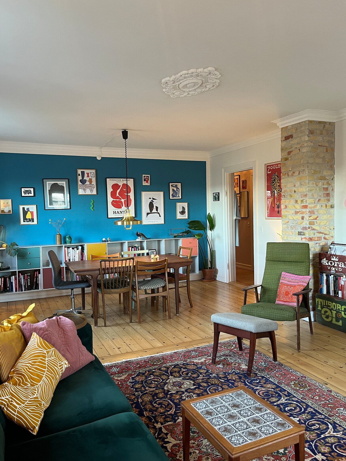 A cozy living space is showcased, featuring a green sofa adorned with vibrant cushions. A wooden dining table with chairs sits at the center, surrounded by colorful wall art. Natural light brightens the area, with a patterned rug on the hardwood floor adding warmth.