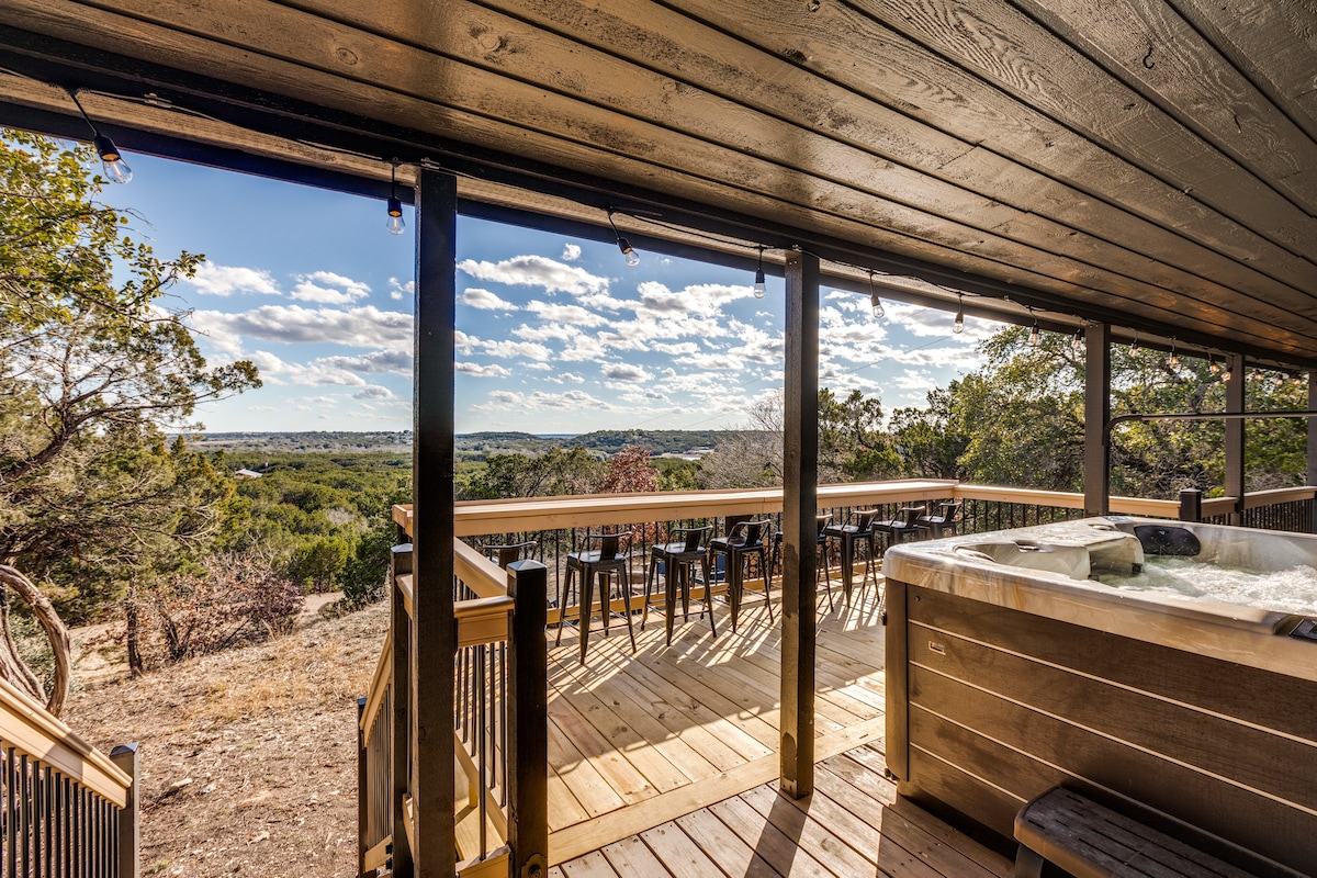A spacious deck features a hot tub positioned for scenic views of the surrounding landscape. Outdoor seating is available nearby, with a row of chairs lining the deck. Large windows allow natural light to fill the space, enhancing the overall ambiance.