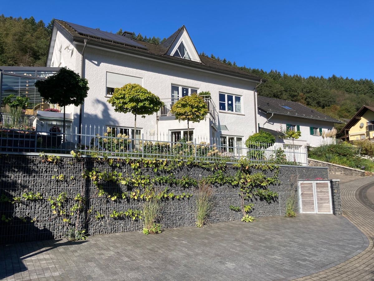 The exterior of the holiday apartment is captured under clear blue skies, highlighting a white two-story building. Vertical gardens and neatly trimmed trees frame the entrance, with a paved driveway leading up to the property. Surrounding greenery and residential homes are visible in the background.