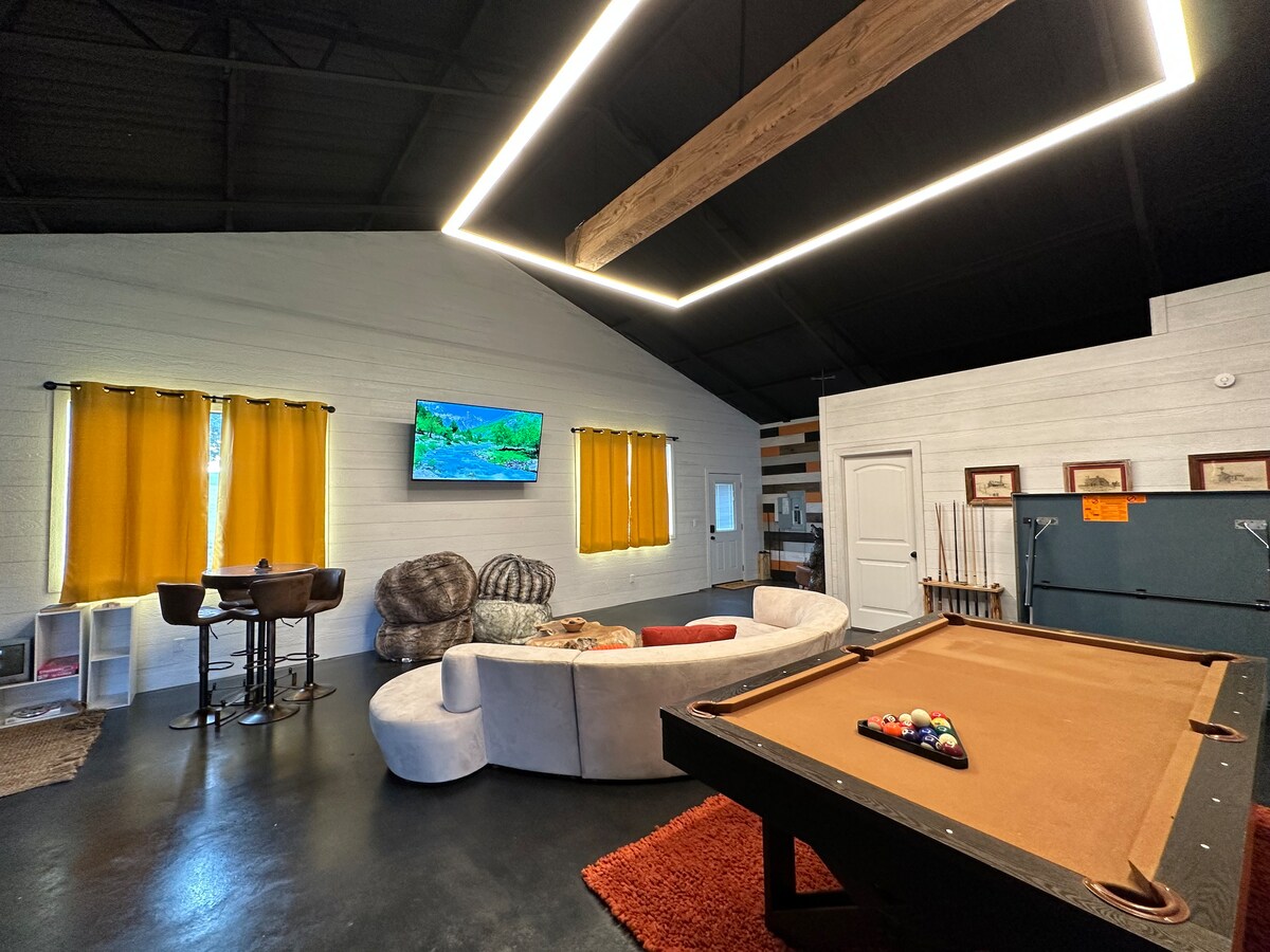 An open living area features a large sectional sofa and a billiards table. A flat-screen television is mounted on the wall, with bright yellow curtains framing the windows. A small dining set is positioned nearby, enhancing the communal feel of the space.