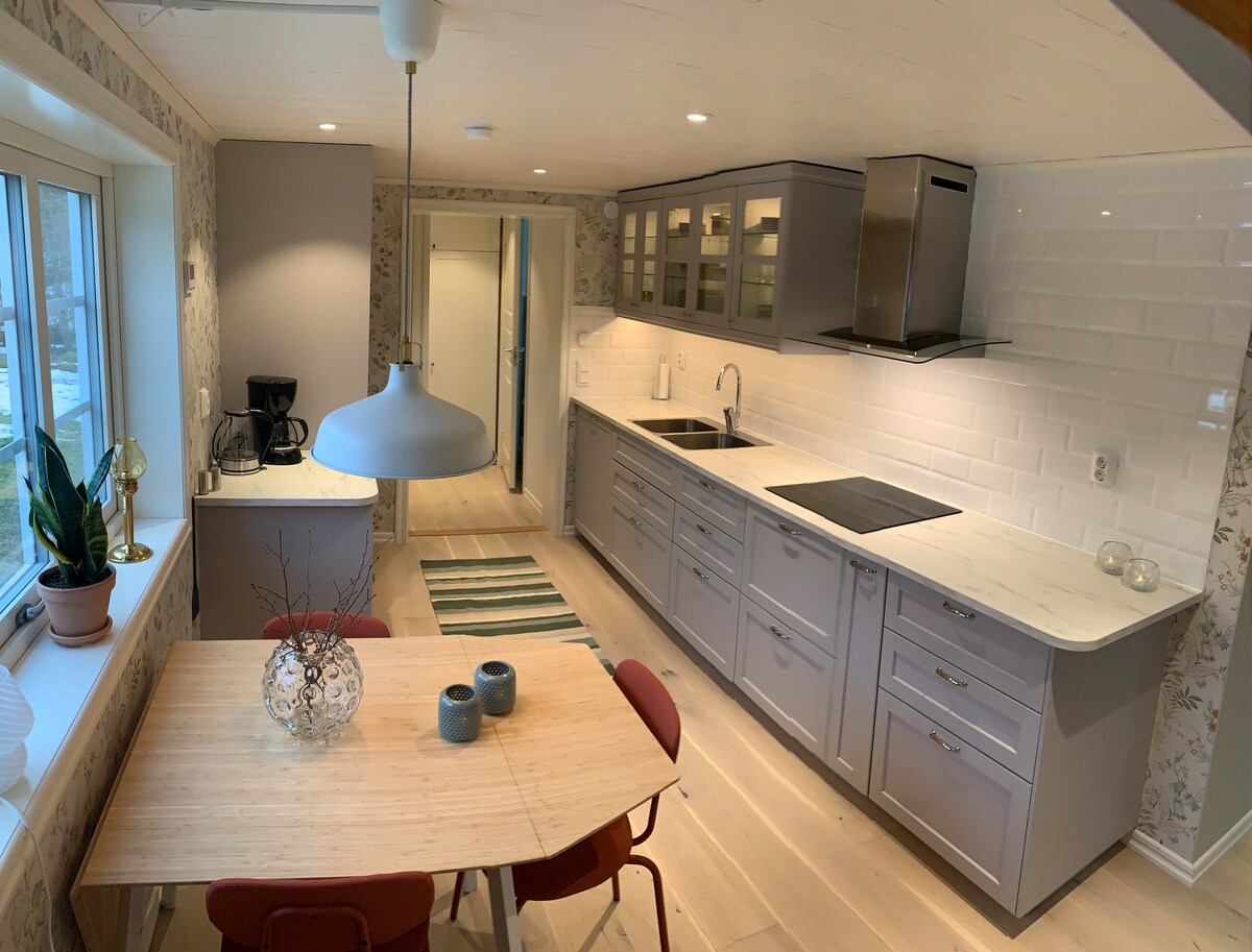 The kitchen features modern cabinets in soft gray, complemented by a marble countertop. A round dining table with red chairs is positioned near a window, providing natural light. Cooking supplies, such as a coffee maker and stovetop, are visibly organized within the space.