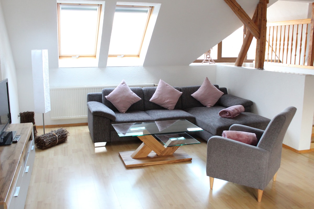 A spacious living area features a comfortable sectional sofa adorned with decorative pillows, paired with a glass coffee table. Large skylight windows allow natural light to fill the room, highlighting the warm wooden accents and sleek television unit.