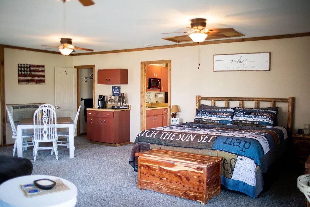 A spacious interior features a comfortable bed adorned with a patterned quilt. A wooden trunk sits at the foot of the bed, while a dining area with a white table and chairs is visible. The kitchenette includes cabinets, a microwave, and coffee supplies.