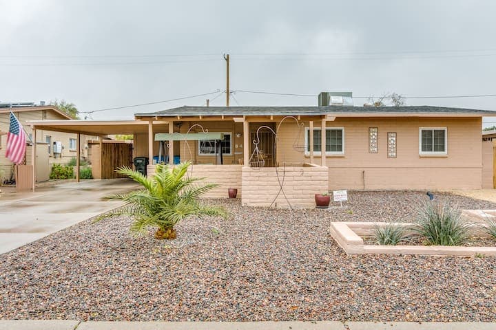 The front exterior of a mid-century single-family home is shown, featuring a covered porch with a hanging swing. A landscaped gravel yard includes a small palm tree and potted plants. An American flag is displayed, and a carport is visible on the side of the home.