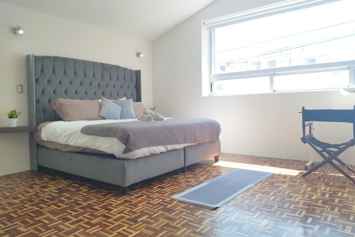 A spacious sleeping area is highlighted by a tufted bed with soft bedding and accent pillows. Natural light floods through the large window, illuminating the unique patterned floor. A small decorative plant sits on the bedside table, while a foldable chair is positioned nearby.