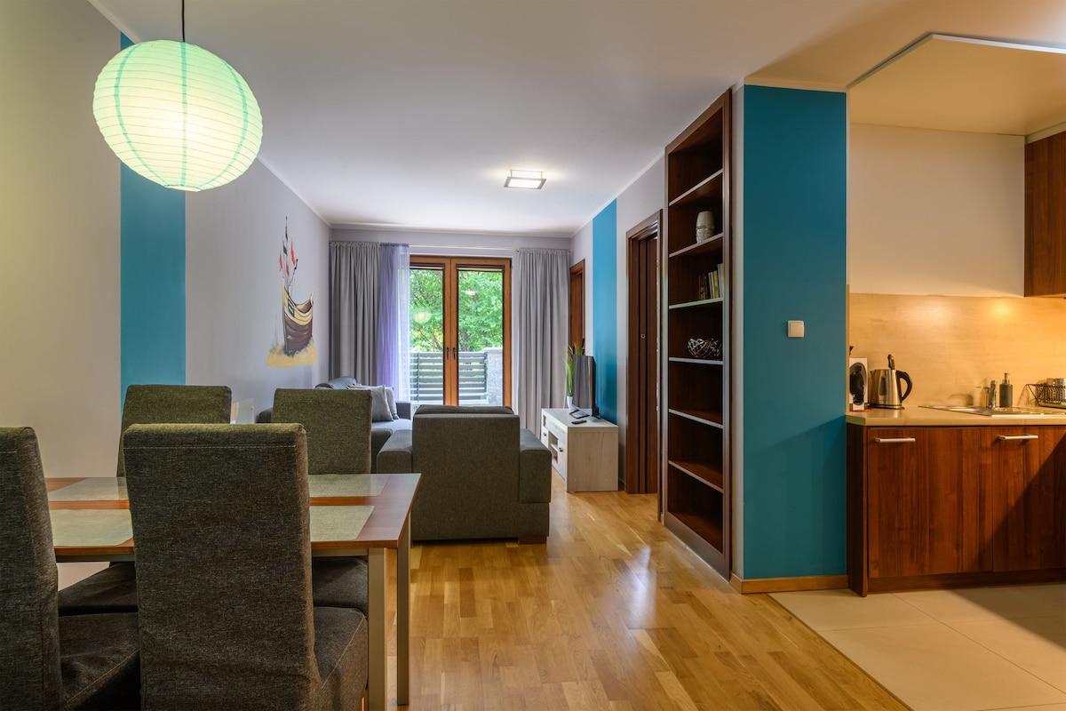 A living area showcases a dining table with four chairs, next to a spacious sofa and a flat-screen TV. Light wood flooring complements the blue and neutral walls. A kitchen area is visible, featuring wooden cabinetry and modern appliances. Natural light enters through large sliding doors.