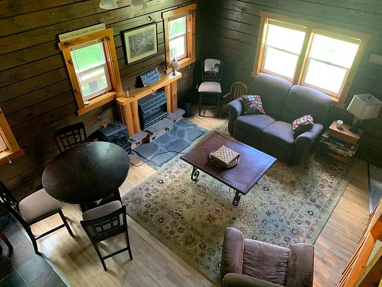 The living area features wooden walls and large windows providing natural light. A comfortable sofa and armchair are positioned around a coffee table on a patterned area rug. A dining table is present, with four chairs arranged around it. A decorative fireplace is also visible.