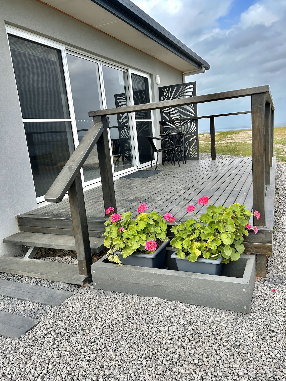 A wooden deck features a railing leading to a well-lit entrance. Potted pink geraniums can be seen on the side, adding a touch of color. The background includes large windows with a scenic view, while decorative panels provide privacy.