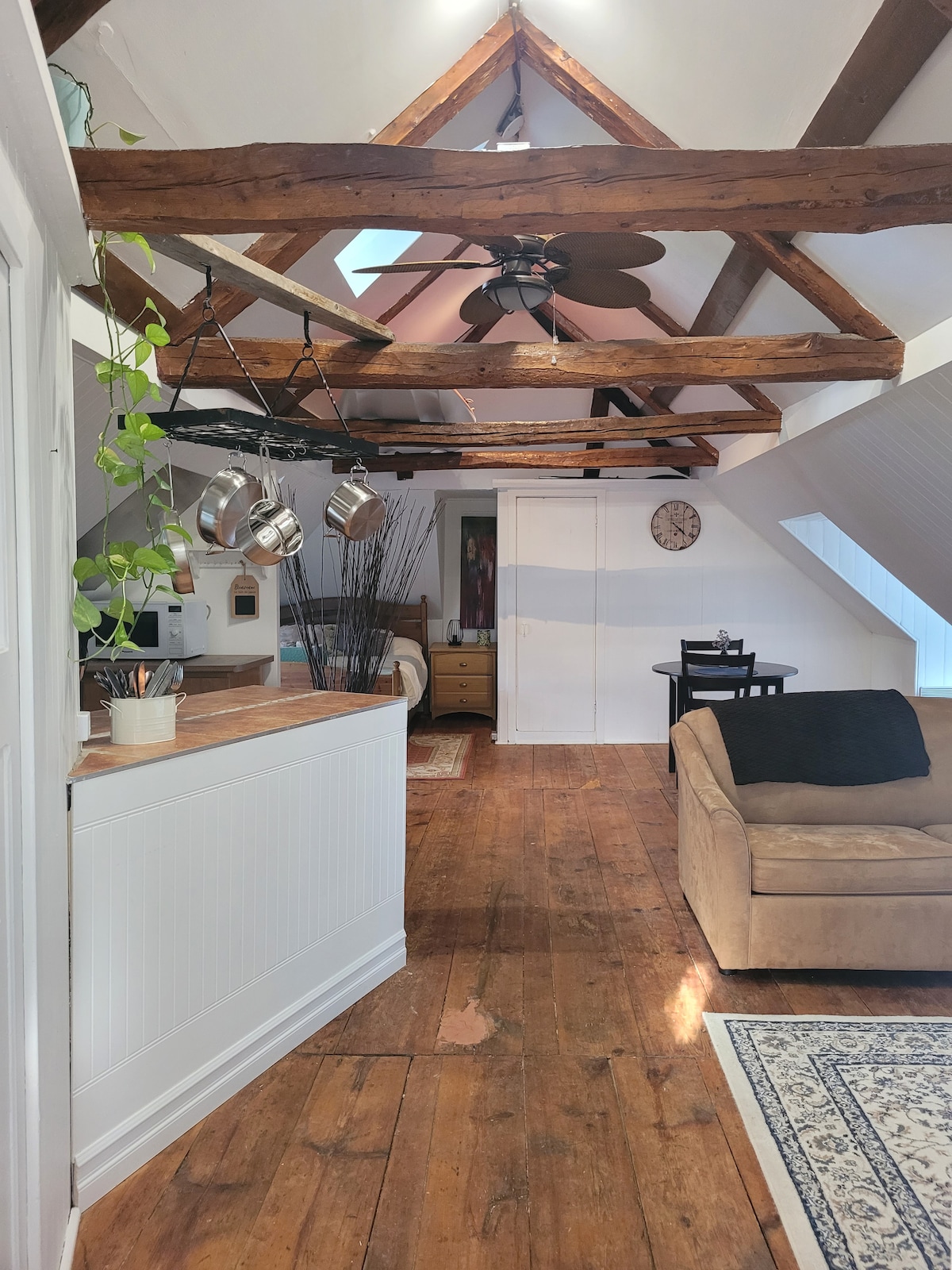 The open loft features a cozy living area with a beige sofa, wooden flooring, and exposed beams overhead. A kitchenette with hanging pots is visible, along with a small dining area and a storage closet. Natural light enters through a window, creating a warm ambiance.