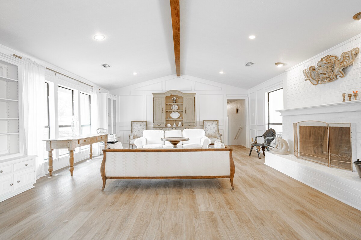 A bright and airy living space features a large white sofa and two armchairs arranged around a central coffee table. Natural light floods the room through large windows, enhancing the light wood flooring. A decorative fireplace is positioned against a white brick wall, adding to the charming ambiance.