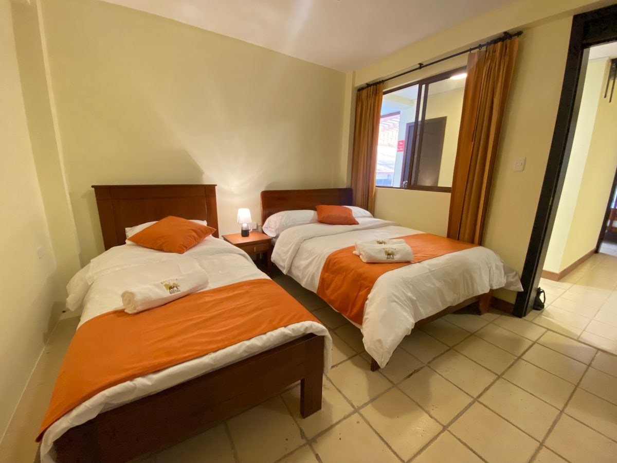 A well-lit room features two beds, one with an orange accent blanket and the other with a white blanket. A nightstand with a lamp is positioned between the beds. Sunlight filters through the curtains, highlighting the neutral-colored walls.