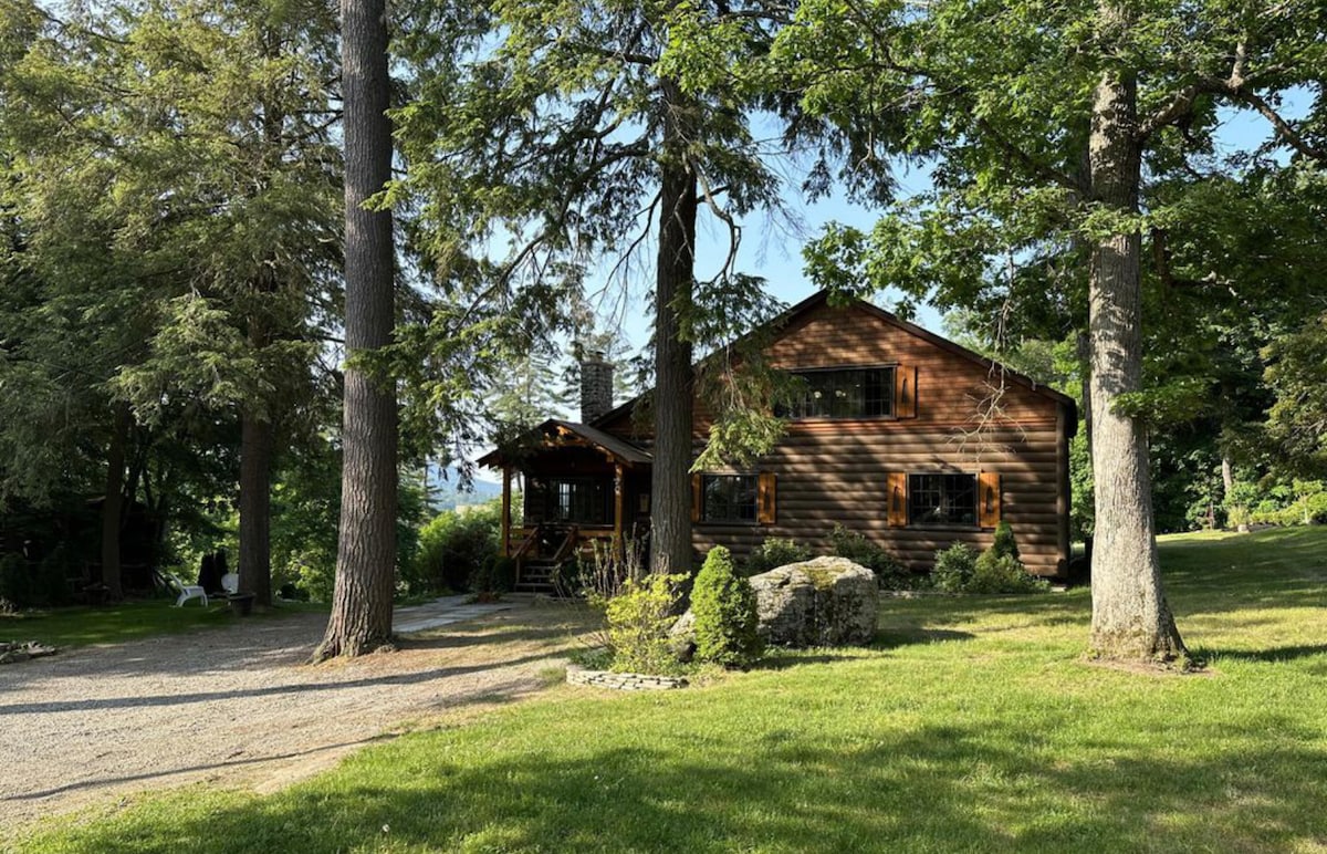 A newly renovated log cabin is surrounded by trees, showcasing a spacious exterior with wooden siding and multiple windows. A gravel path leads to the entrance, while a lush green lawn lies in front, providing a serene setting for relaxation and outdoor activities.