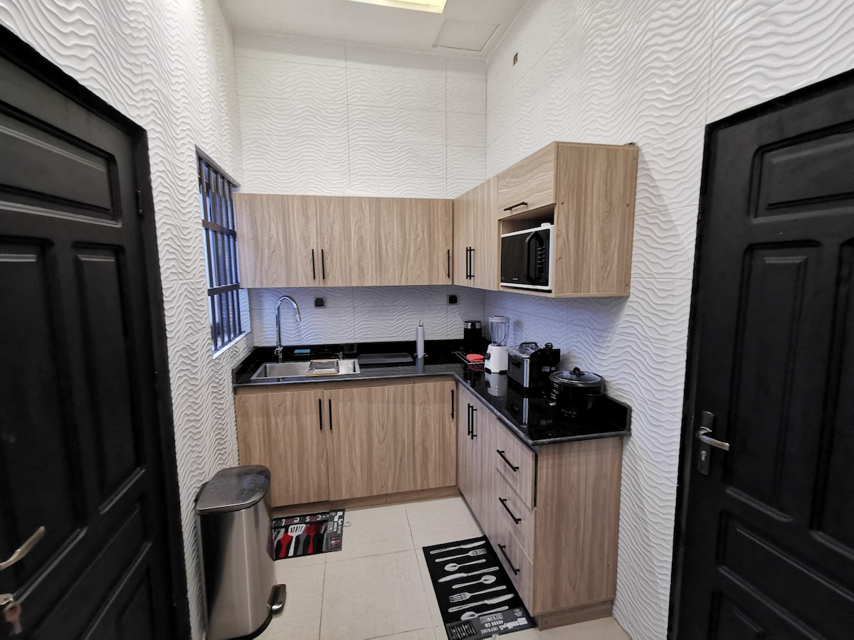 A modern kitchen is presented with light wood cabinetry and black countertops. Stainless steel appliances and a microwave are visible, along with a sink integrated into the counter. The space is well-lit, featuring textured walls and tiled flooring.