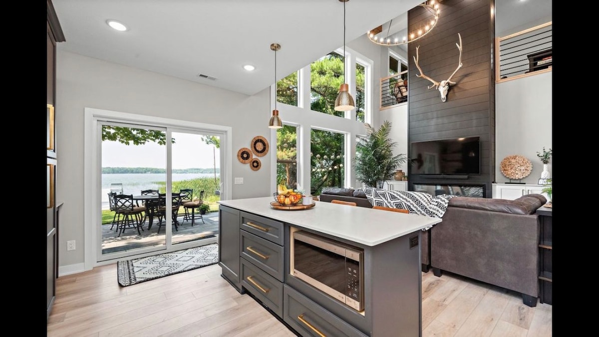 A modern kitchen features an island with a built-in microwave and pendant lighting. Large windows frame lake views, while a cozy seating area with a fireplace is visible. A dining set is arranged on an outdoor patio, surrounded by greenery.