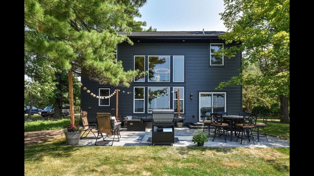 A spacious backyard is shown, featuring comfortable seating arrangements and a dining table. Large windows offer views of the interior. Decorative string lights are displayed above the seating area, and greenery surrounds the space, adding a natural element to the inviting outdoor setting.