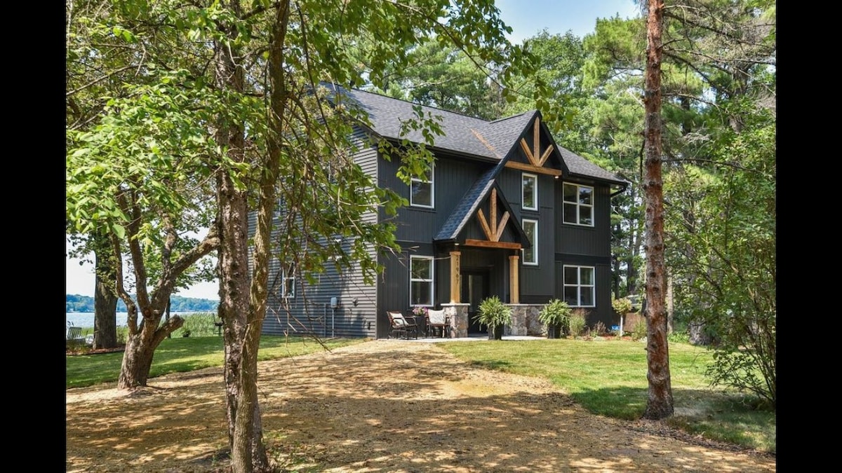 A modern two-story home, framed by trees, features a spacious front porch and multiple windows. The exterior is adorned with a mix of dark siding and natural wood accents. A manicured lawn leads to a serene lake view in the distance.