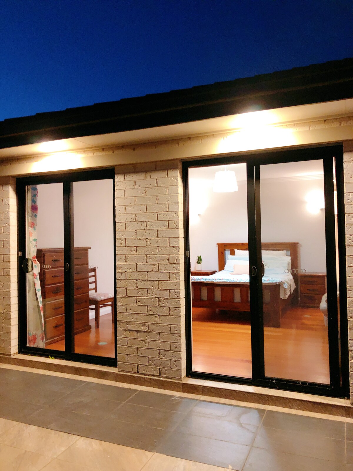 A view of a well-lit bedroom through expansive glass doors, showcasing a spacious bed with matching bedside tables. The warm wooden floors contrast with the light walls, while soft lighting enhances the room's inviting ambiance.