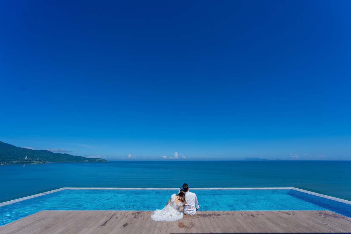 The infinity pool stretches along the edge of the terrace, offering uninterrupted views of the sea and sky. The calm waters reflect the expansive blue above, while gentle waves can be seen in the distance. A couple is seated nearby, enjoying the serene atmosphere.