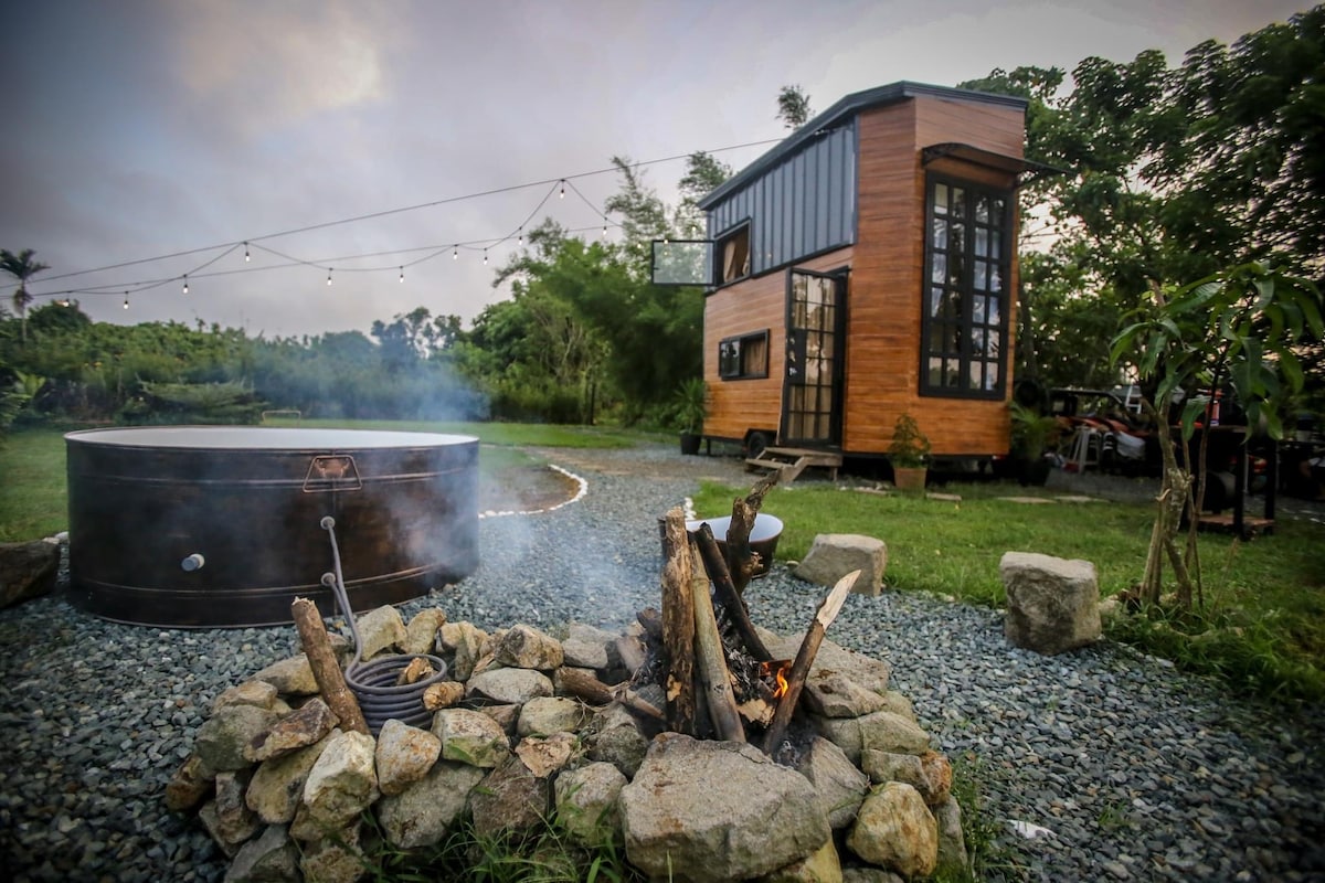 An outdoor scene featuring a tiny house with a wooden exterior and large windows. Nearby, a circular outdoor tub is situated alongside a stone fire pit with a small fire burning. String lights are seen above, enhancing the tranquil ambiance.