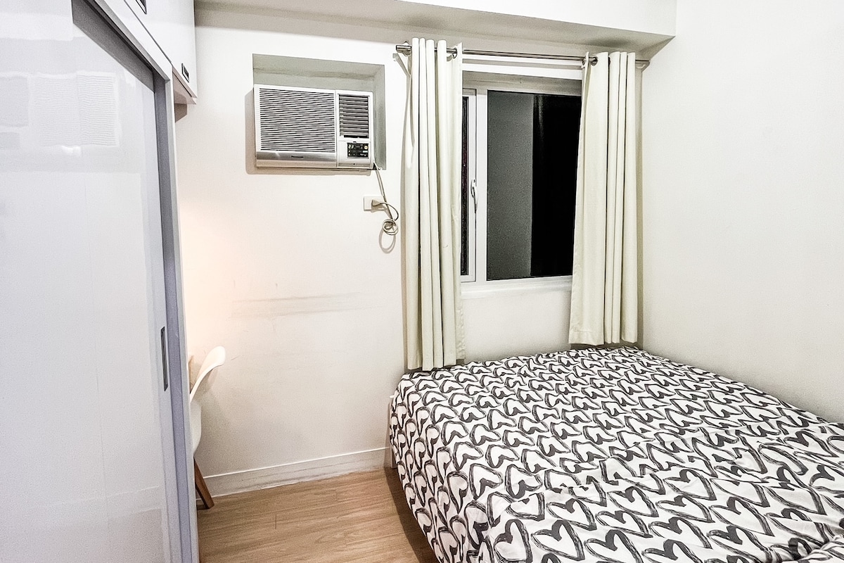 A cozy bedroom features a queen-size bed with a patterned duvet. An air conditioning unit is mounted above the window, which is dressed with curtains. A closet cabinet is visible to the left, and a chair is positioned nearby.