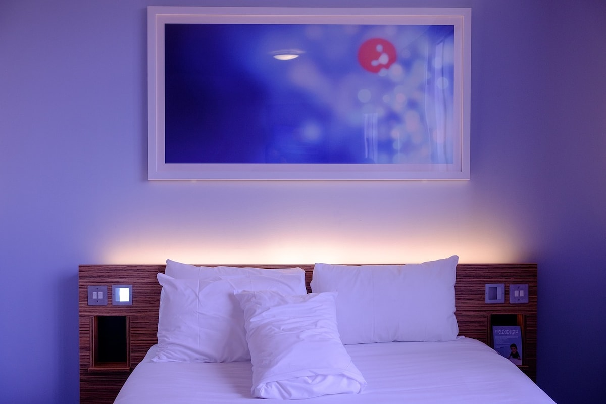 A comfortable bed with plush white bedding is centered in the room, complemented by a soft glow from integrated lighting on the headboard. Above, a framed artwork adds a subtle pop of color against the calming blue walls, creating a serene atmosphere.