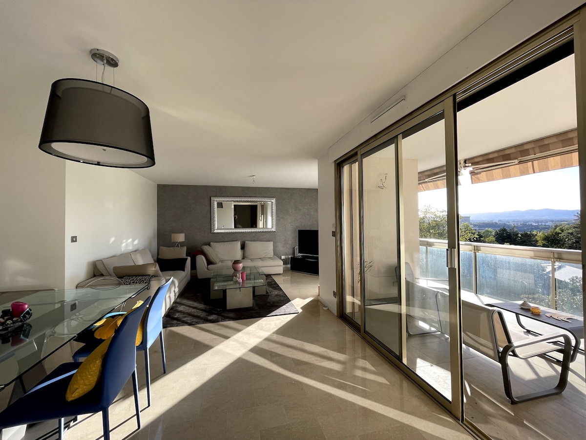 A spacious living area features large glass doors leading to a terrace, allowing natural light to enter the room. A comfortable sectional sofa and a flat-screen TV are present. A dining table with colorful chairs is positioned nearby, complementing the modern decor and scenic views.