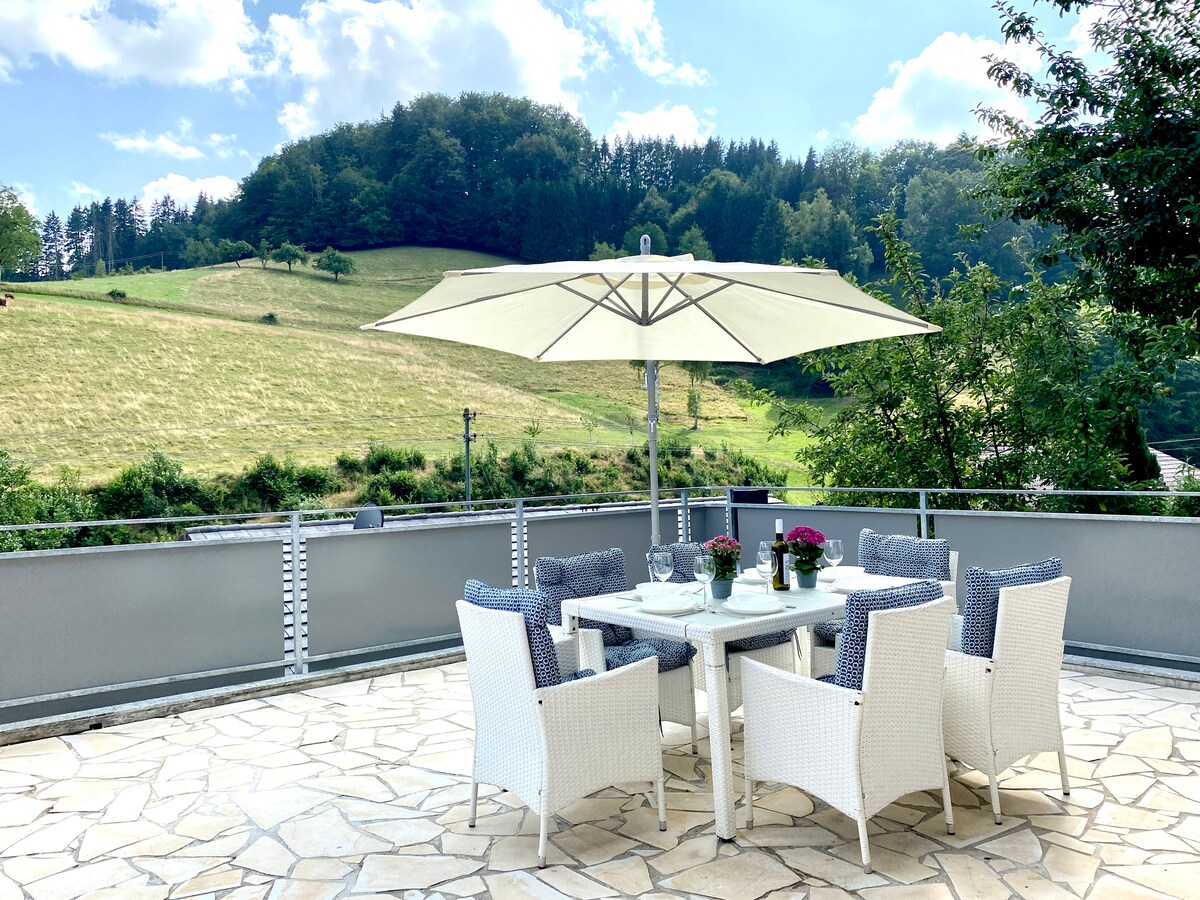 A spacious terrace features a large table surrounded by six wicker chairs. An umbrella provides shade over the table, which is adorned with potted flowers. The backdrop showcases a scenic view of rolling hills and trees under a bright blue sky.