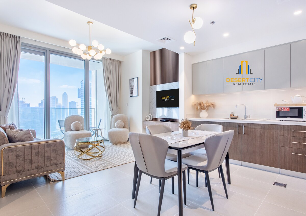 An open-plan living and dining area showcases modern furniture, including a round dining table with four upholstered chairs. A plush sofa and a stylish accent chair are positioned near large floor-to-ceiling windows that frame a view of the skyline.
