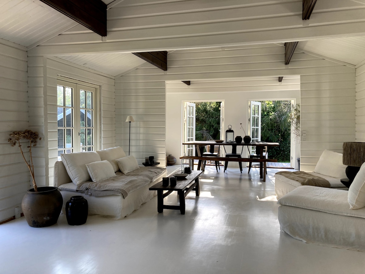 The open living space features a bright, airy atmosphere with large windows allowing natural light. Comfortable seating is arranged with neutral-colored cushions, while a rustic wooden table sits center in the room. A minimalist decor style creates a relaxing environment.