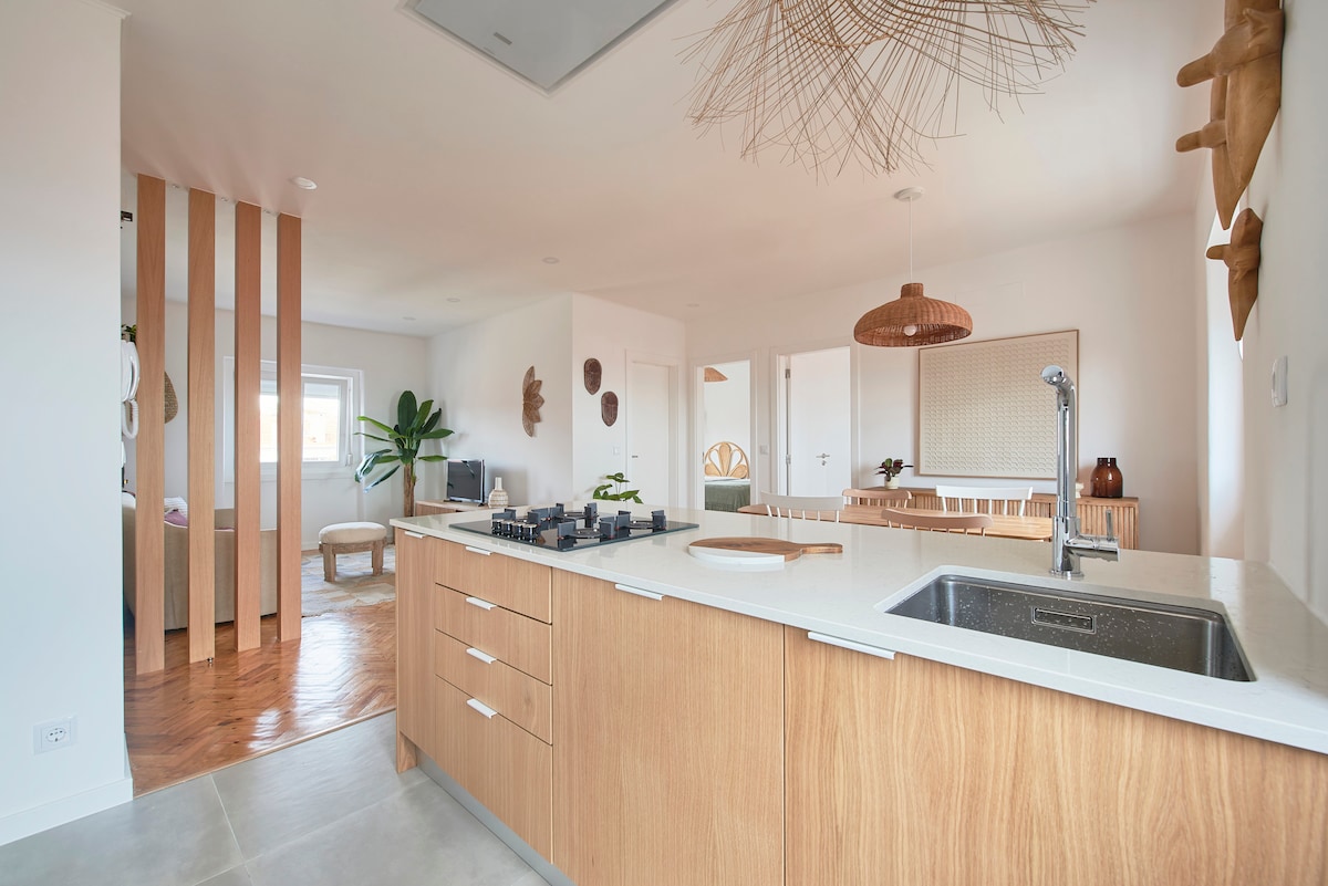 An open-concept kitchen and living area is displayed, featuring a modern kitchen island with a sink and stovetop. Natural wood cabinetry contrasts against white countertops. Light wood accents and plants are visible, creating a bright, airy atmosphere.