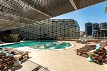 An outdoor pool area features a distinct geometric structure overhead, providing shade. The pool is surrounded by several lounge chairs, while a hot tub is positioned to one side, with city buildings visible in the background.