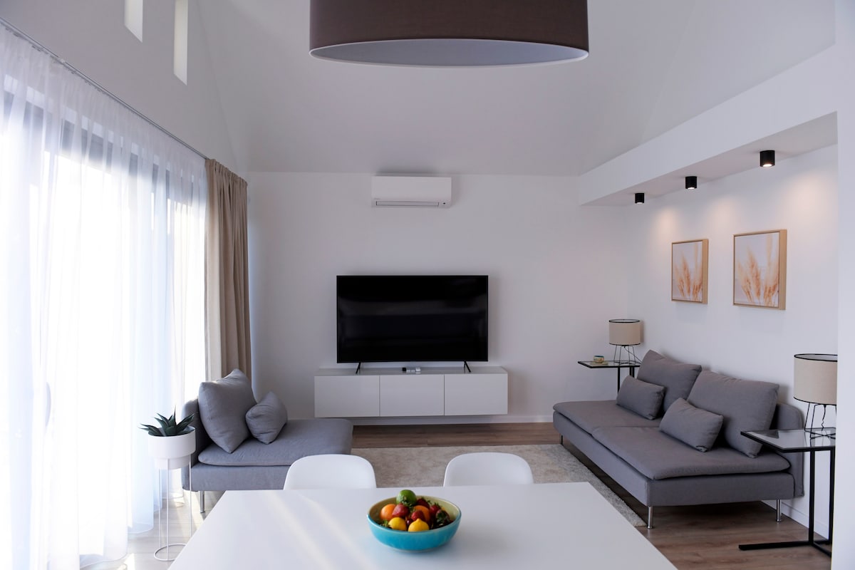 A spacious living area features a modern sectional sofa positioned next to a sleek entertainment unit. A dining table is set with a bowl of fresh fruits. Natural light filters through sheer curtains, creating a bright and airy atmosphere, complemented by stylish wall decor.