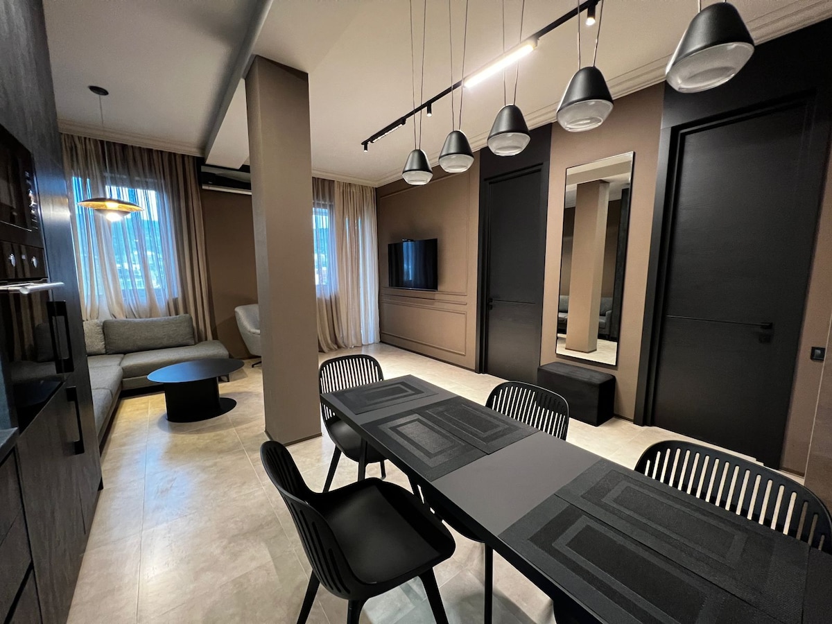 A modern living area is highlighted by a sleek dining table surrounded by four chairs. A comfortable seating arrangement includes a sofa and armchair. Large windows are adorned with soft drapes, and a flat-screen TV is mounted on the wall. Warm tones and ambient lighting create a cohesive space.