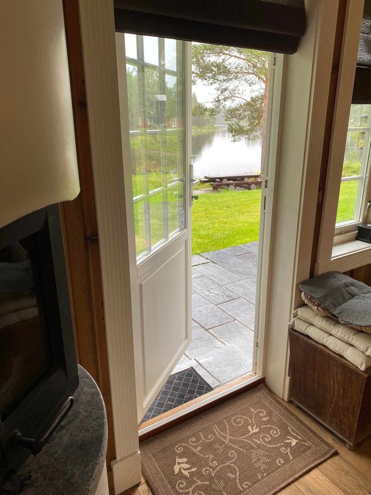 A doorway with a white frame is open, revealing a view of lush green grass and a serene water surface. A stone path leads outside, complemented by a doormat positioned inside the entrance. The interior features a cozy seating area beside a fireplace.