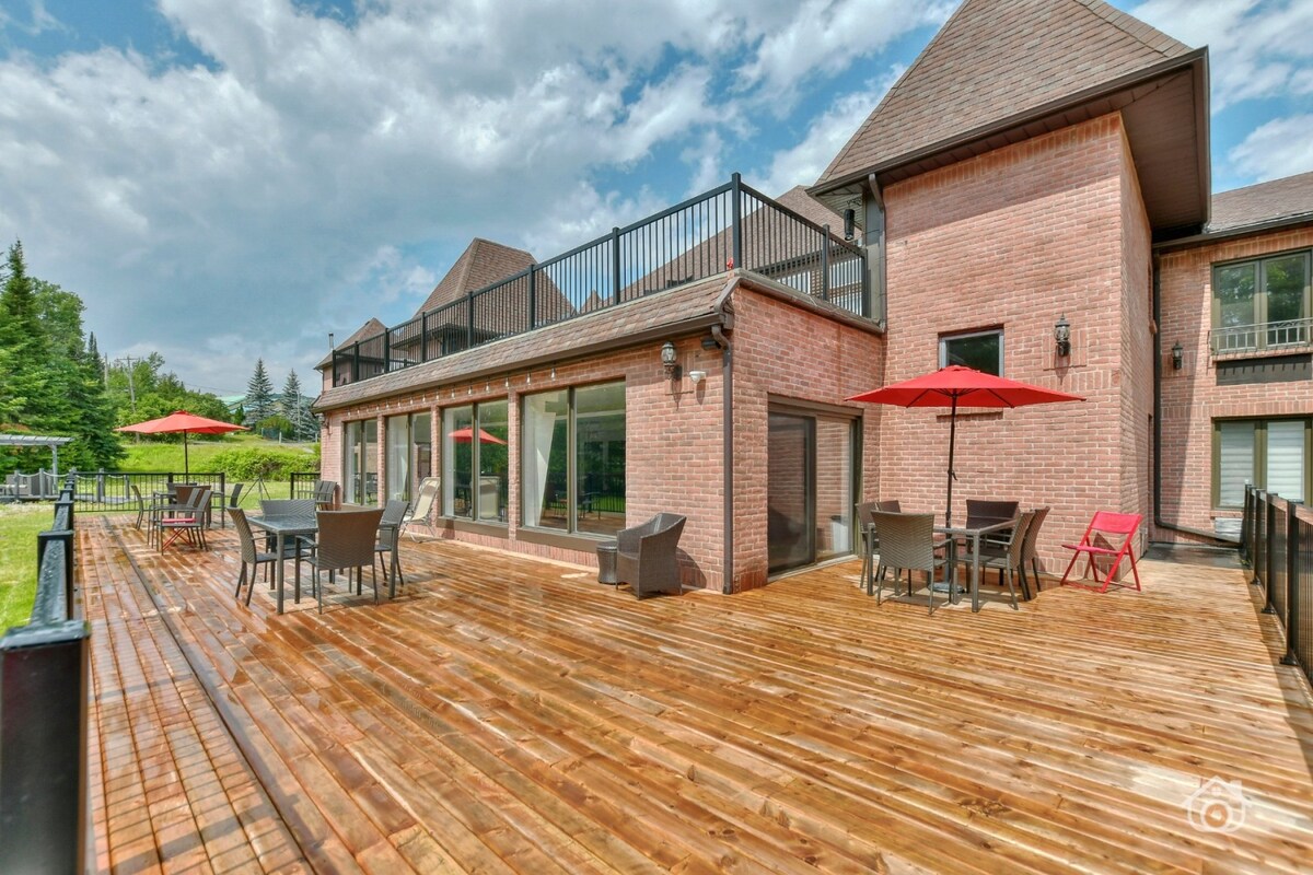 A spacious wooden deck features outdoor seating arrangements, including tables and umbrellas. The area is bordered by lush greenery, providing a serene backdrop. Large glass windows from the attached building invite natural light, enhancing the inviting atmosphere of the outdoor space.