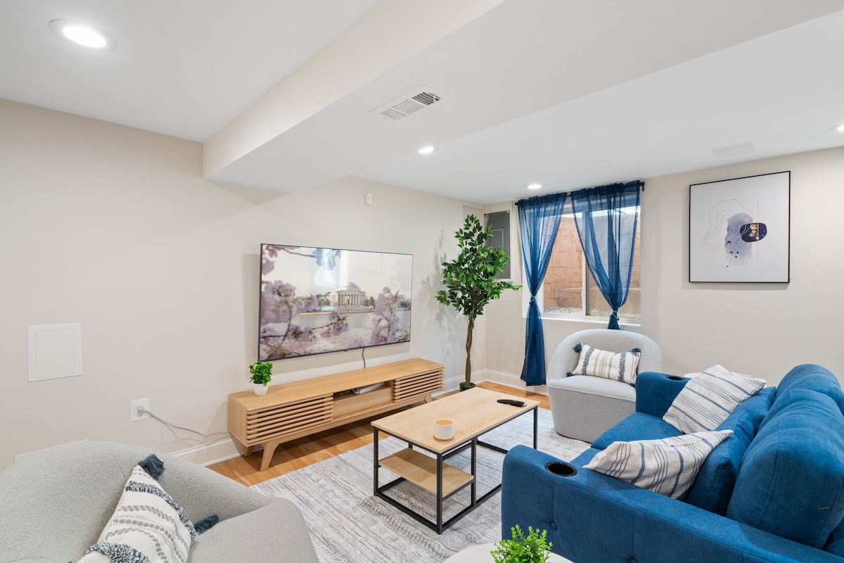 The entertainment area showcases a spacious layout with a large Smart TV mounted on a light wooden media console. Soft seating is provided by a blue sofa and two armchairs. Natural light enters through a window adorned with flowing curtains, while a coffee table sits centrally, inviting relaxation.