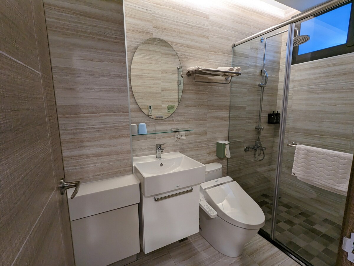 A modern bathroom is presented with a light-colored tiled wall and floor. A sleek vanity with a round mirror is located above a white sink. A walk-in shower with glass panels is featured, along with a heated towel rail and a wall-mounted toilet.