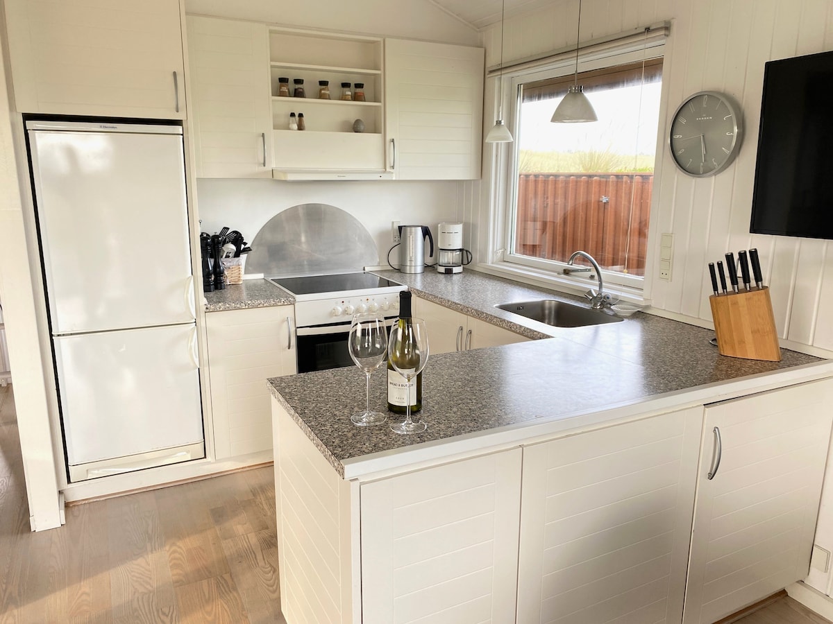 A modern kitchen is equipped with white cabinetry and a spacious countertop. Stainless steel appliances, including a refrigerator and a sink, are visible. A set of wine glasses and a bottle of wine are placed on the counter, accompanied by a knife block and a clock on the wall.