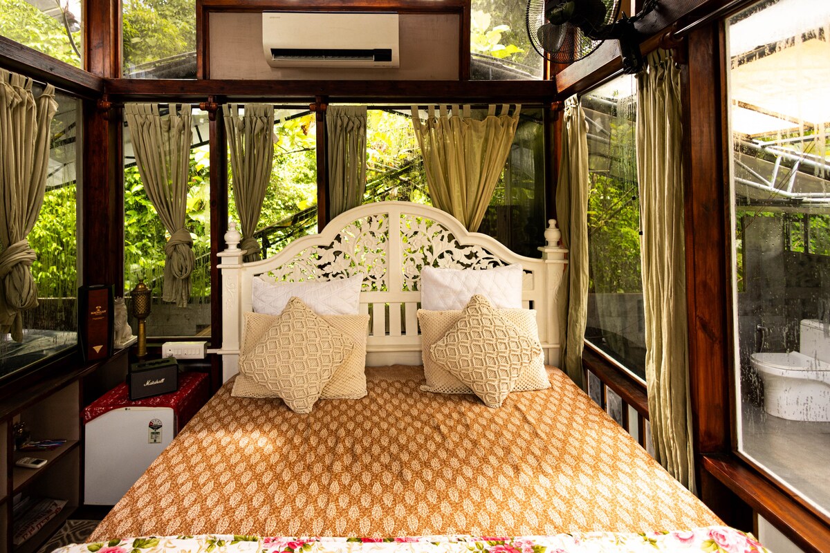 A spacious bed is centered in a glass room, featuring decorative pillows and a patterned blanket. Natural light filters in through lush greenery outside, enhancing the serene environment. Nearby, a small nightstand and a modern air conditioner are visible, contributing to the room's functionality.