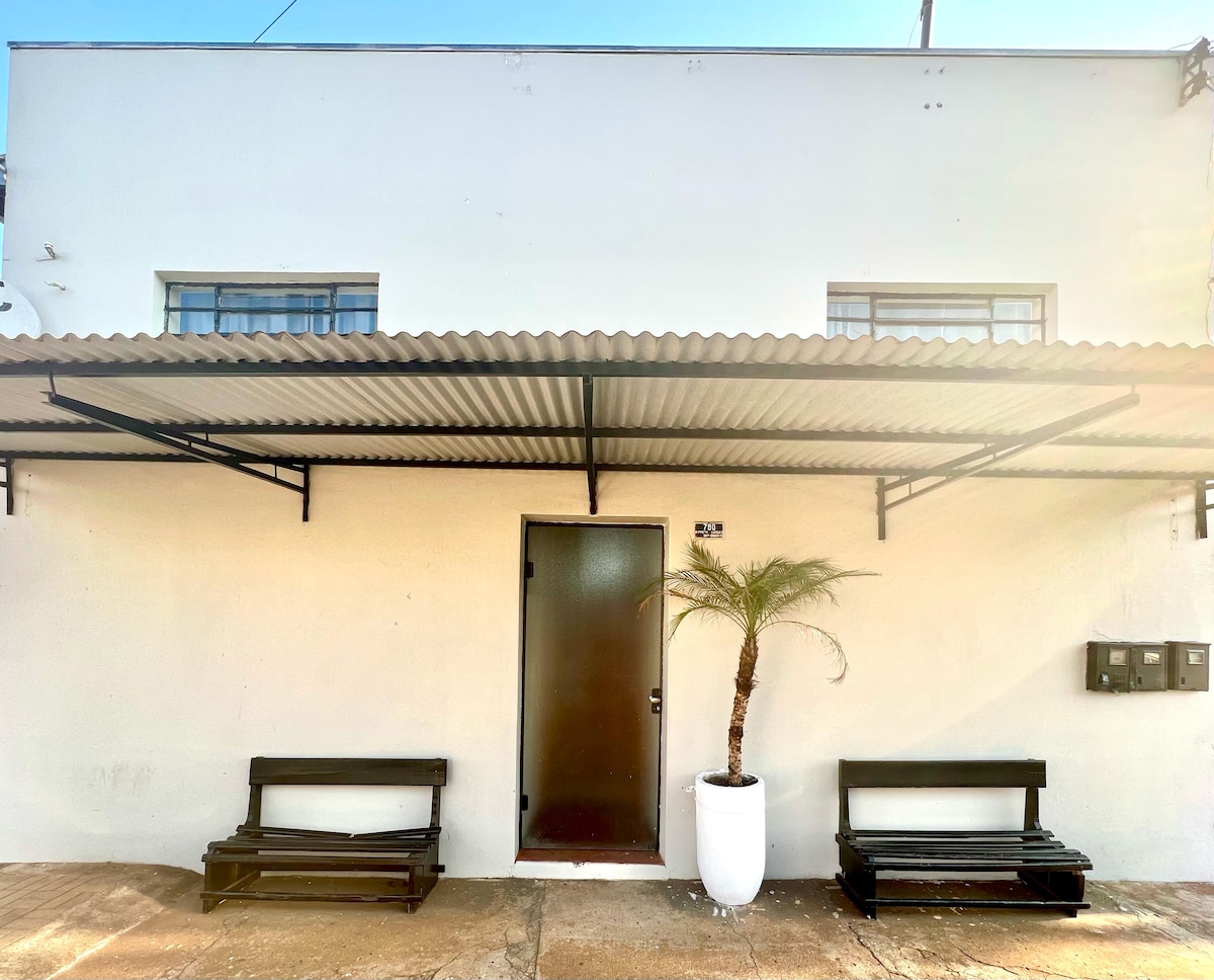 The exterior of the house is showcased, featuring a plain white facade with a dark entrance door. Two black benches are positioned on either side of the entrance beneath a covered awning. A potted palm adds a touch of greenery, enhancing the welcoming appearance.