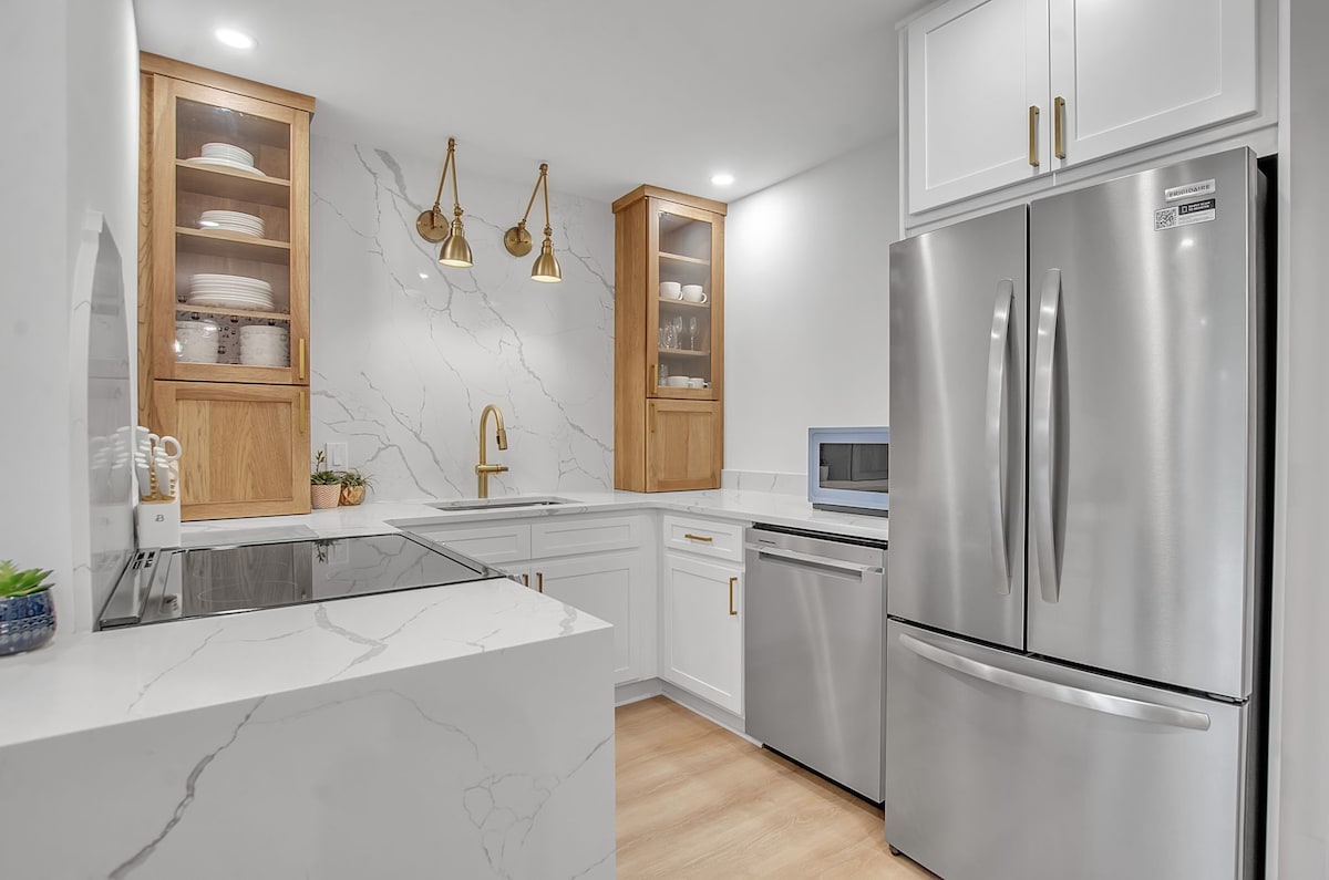 A modern galley kitchen features sleek stainless steel appliances, including a refrigerator and microwave. Shaker cabinets with glass doors display decorative dishware. A spacious countertop extends from the left, complemented by a gold-finished faucet and subtle lighting above, creating an inviting cooking space.