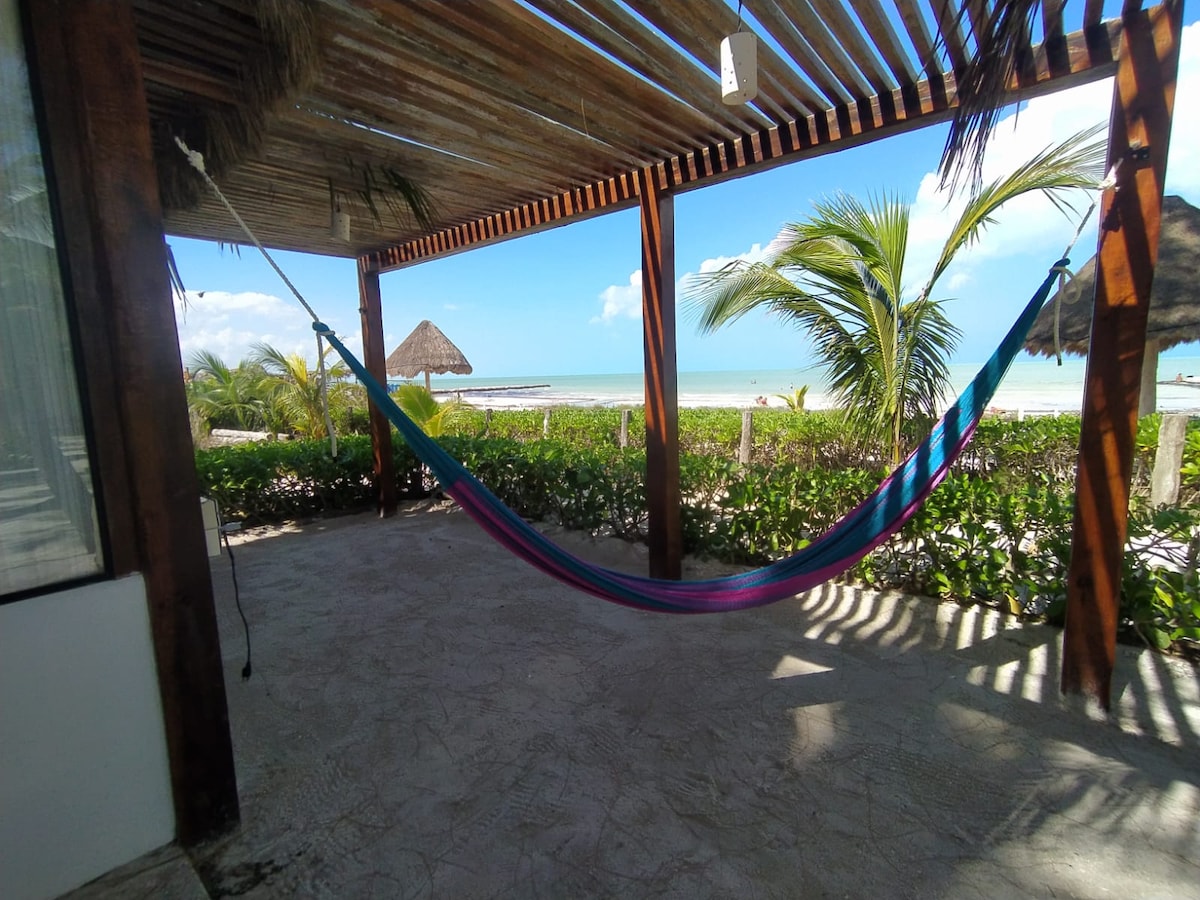 A colorful hammock is suspended under a wooden pergola, offering a serene view of the beach and ocean. Lush greenery lines the patio, with palm trees swaying gently in the breeze. A thatched umbrella can be seen in the distance against a clear sky.