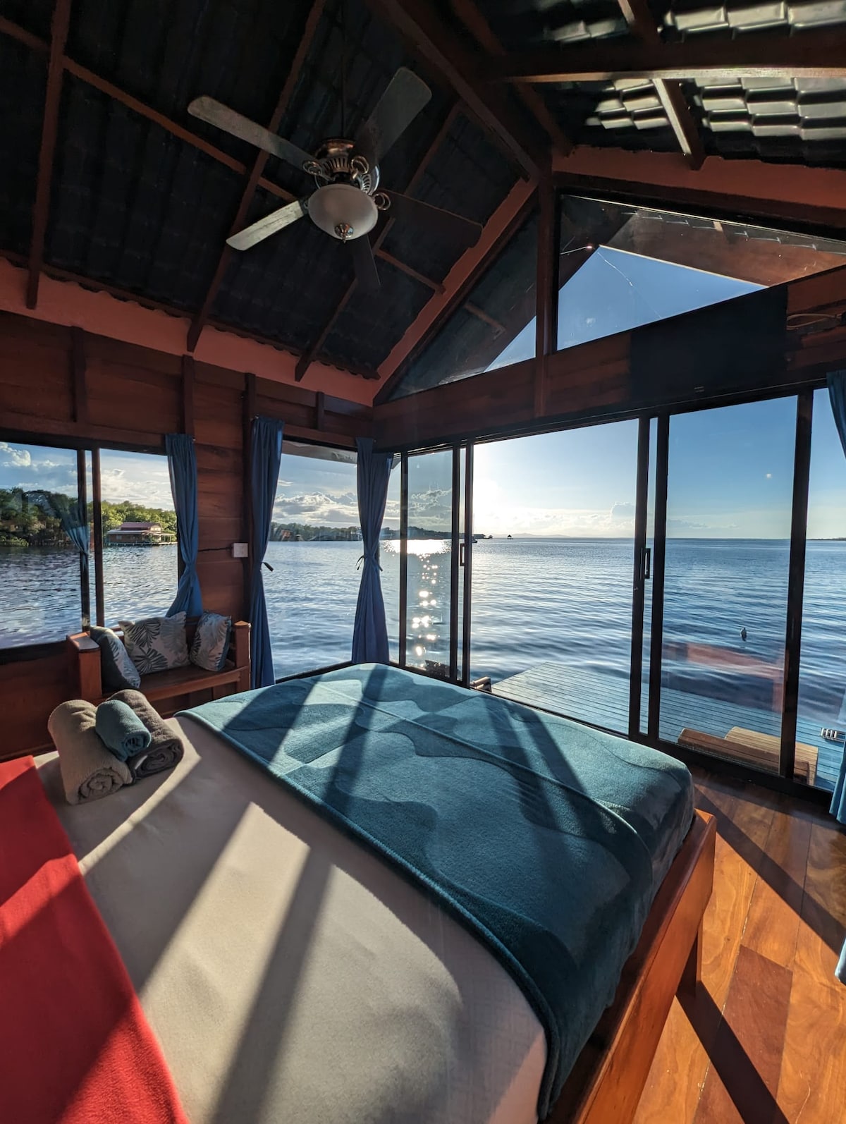 A spacious bedroom features large glass doors offering views of the tranquil waters. Natural light filters through, casting serene patterns on the wooden floor. The inviting bed is adorned with a soft blue blanket, with a ceiling fan visible above.