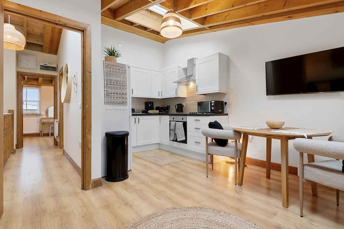 The kitchen is fully equipped with white cabinetry, a stove, refrigerator, and ample countertop space. A dining table for four is positioned near a wall-mounted TV. Natural light is provided by a skylight above, highlighting the wooden ceiling beams.