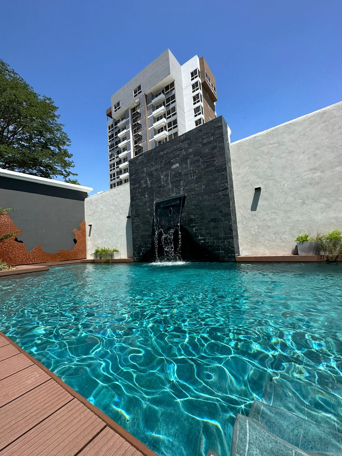 A serene pool area is showcased, featuring clear turquoise water with gentle ripples. A modern stone wall with a water feature adds a calming element, while a tall apartment building stands in the background against a bright blue sky.