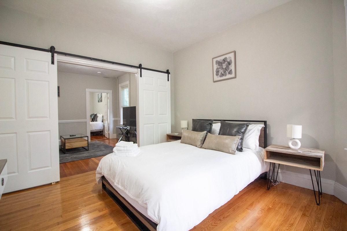 A cozy bedroom features a spacious bed with a white duvet and decorative pillows. The room is illuminated by natural light from a nearby window. Two bedside tables with lamps are placed on either side of the bed, contributing to a clean and simple design.